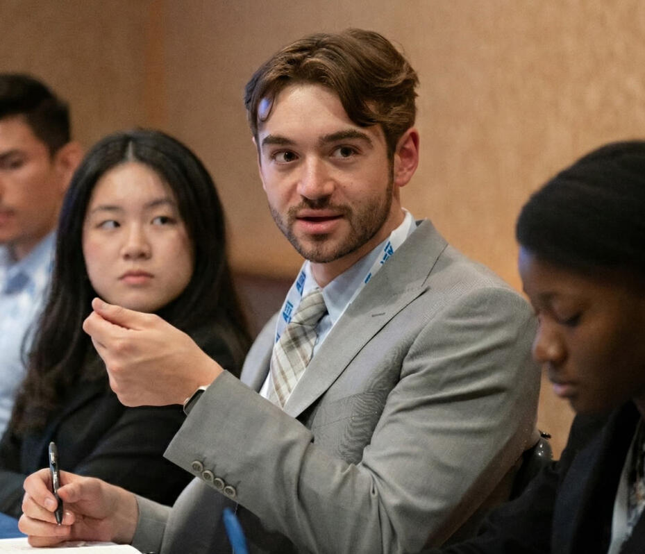 Micah DenBraber speaking on a conference panel, gesturing with a pen in hand.